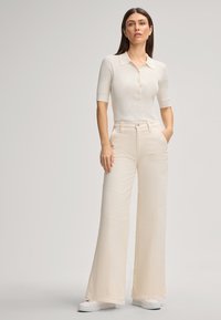 Donna in maglietta a polo lavorata a maglia crema, pantaloni a vita alta e gamba larga crema e sneaker bianche contro uno sfondo chiaro e semplice.