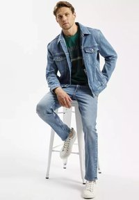 Jasnoniebieska kurtka dżinsowa i dżinsy, z zieloną koszulką pod spodem. Białe trampki z czerwonymi akcentami. Model siedzi na stołku.