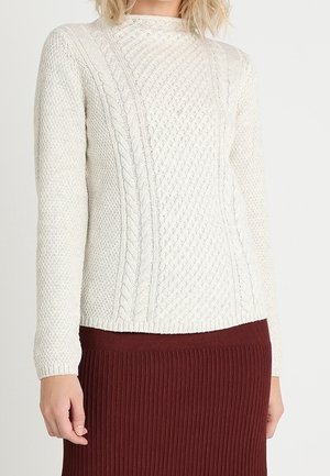 Kobieta ubrana w kremowy sweter warkoczowy z długimi rękawami i prążkowaną bordową spódnicę, stojąca na tle jednolitego tła.