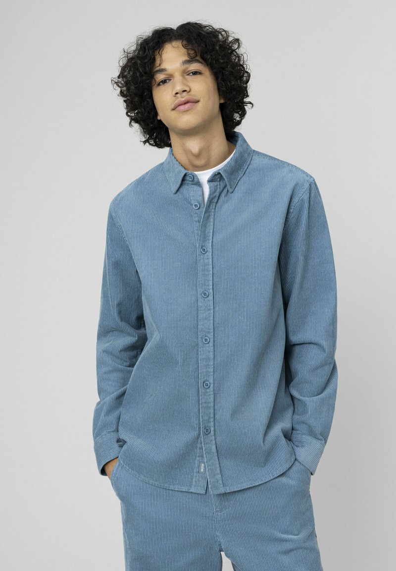 Chemise en velours côtelé bleu clair à boutons, avec col, manches longues et coupe droite, présentant une texture côtelée verticale et des poches avant.