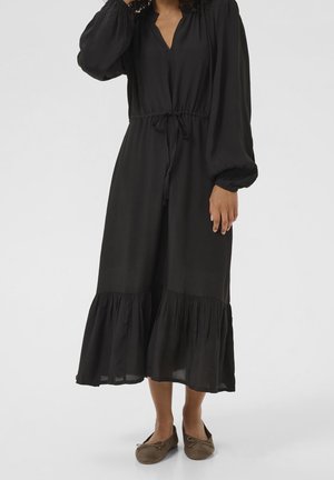 Femme portant une robe midi noire à manches longues avec ourlet froncé et ballerines marron, debout devant un fond uni.