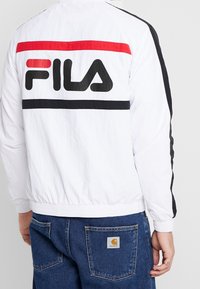 Biały kurtka przeciwwiatrowa z czarnymi i czerwonymi poziomymi paskami, z dużym czarnym logotypem "FILA" z tyłu. Elastyczne mankiety i pas.