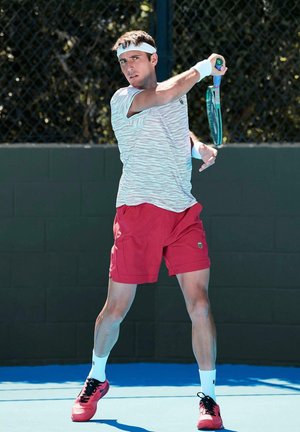 Joueur de tennis masculin en short rouge et t-shirt blanc se préparant à un coup de revers sur un court extérieur avec un revêtement bleu.