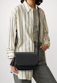Svart läder crossbody-väska med klaffstängning, prydd med en tofs och en lång justerbar rem. Bärs med en randig oversized skjorta.