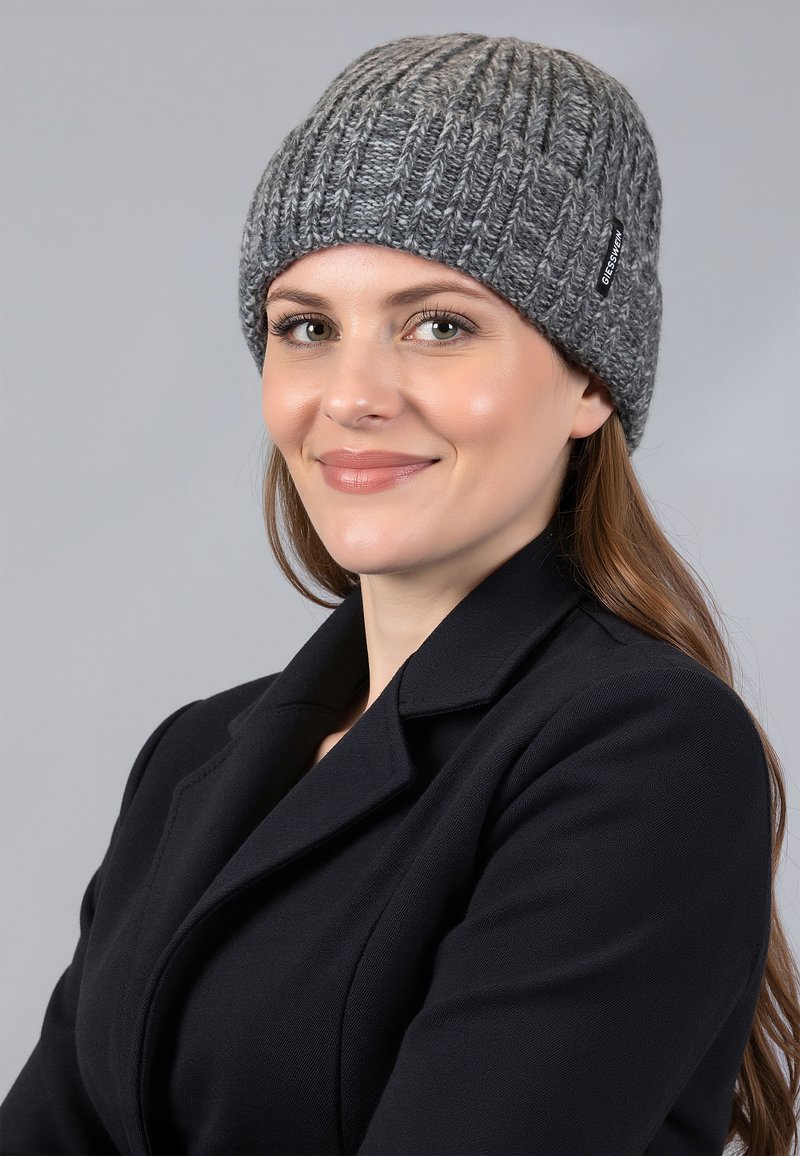 Grijze gebreide beanie met een geribbelde textuur en een klein zwart label. Draag door een persoon in een zwarte jas. Achtergrond is neutraal grijs.