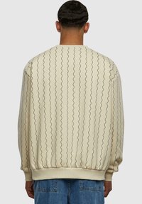 Sudadera beige con líneas negras onduladas, puños y dobladillo acanalados. Presenta un cuello redondo y un ajuste relajado. La tela parece suave y con textura.