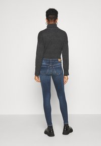 Una persona con cabello corto y rizado se encuentra de espaldas, vistiendo un suéter oscuro, jeans ajustados azules y botas negras sobre un fondo simple.