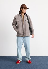 Light grey corduroy jacket with two chest pockets, tan t-shirt underneath, light blue jeans, and red sneakers with white stripes. Brown cap and black bag added.