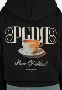 Felpa nera con il grafico di una tazza da caffè su un piattino. Il testo recita "PGDB" e "Peace Of Mind" in caratteri decorativi.