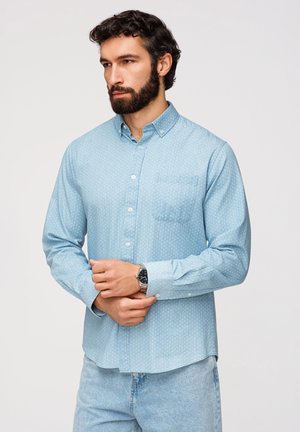 Mężczyzna z ciemnymi włosami i brodą, ubrany w jasnobłękitną koszulę z wzorem i jasnoniebieskie dżinsy, poprawiający swój zegarek.