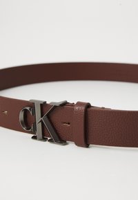 Hnedý kožený opasok s textúrou a čiernou kovovou sponou "CK" ležiaci na bielej ploche.