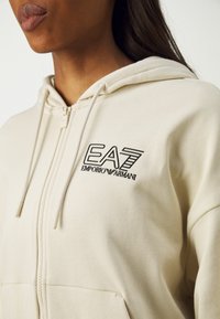 Kremno obarvana zip-up jopica iz mehkega blaga. Ima sprednji žep in črn vezen logotip na prsih. Kapuca z vrvico.
