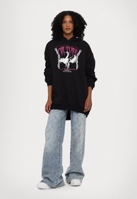 Black sweatshirt with graphic print featuring a swan in white and pink text, paired with light blue wide-leg jeans with logos throughout.