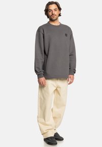 Sudadera gris con cuello redondo, mangas largas y un pequeño parche con logo; combinada con pantalones beige holgados y zapatos negros.