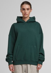 Mørkegrøn oversized hættebluse lavet af blødt stof, med en frontlomme og snoretræk i hætten. Designet har brede ærmer og ribbet manchetter.