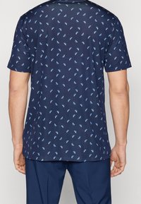 Námořnicky modrá košile s krátkým rukávem a geometrickým vzorem světle modrých trojúhelníků. Vyrobená z texturované látky, má kulatý výstřih a volný střih.