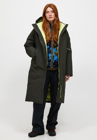 Langer dunkelgrüner Parka mit einer leuchtend gelben Innenkapuze, Frontreißverschluss und Seitentaschen. Über einem gemusterten blauen und braunen Pullover getragen.