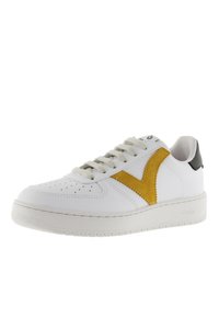 Zapatilla blanca con raya en ante amarillo, cordones en la parte delantera, puntera perforada y tirador negro en la suela blanca.