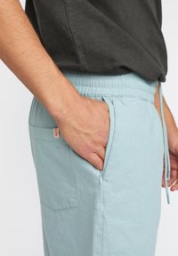 Lichtblauwe, lichte broek met een elastische tailleband, zijzak met rits en een subtiel, geaccentueerd patroon. Gecombineerd met een donkergrijs bovenstuk.
