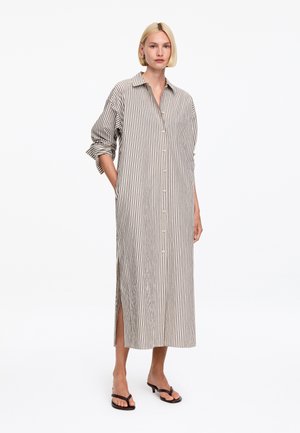 Femme portant une longue robe-chemise ample beige et blanche à rayures verticales, manches retroussées et sandales à talons noires.