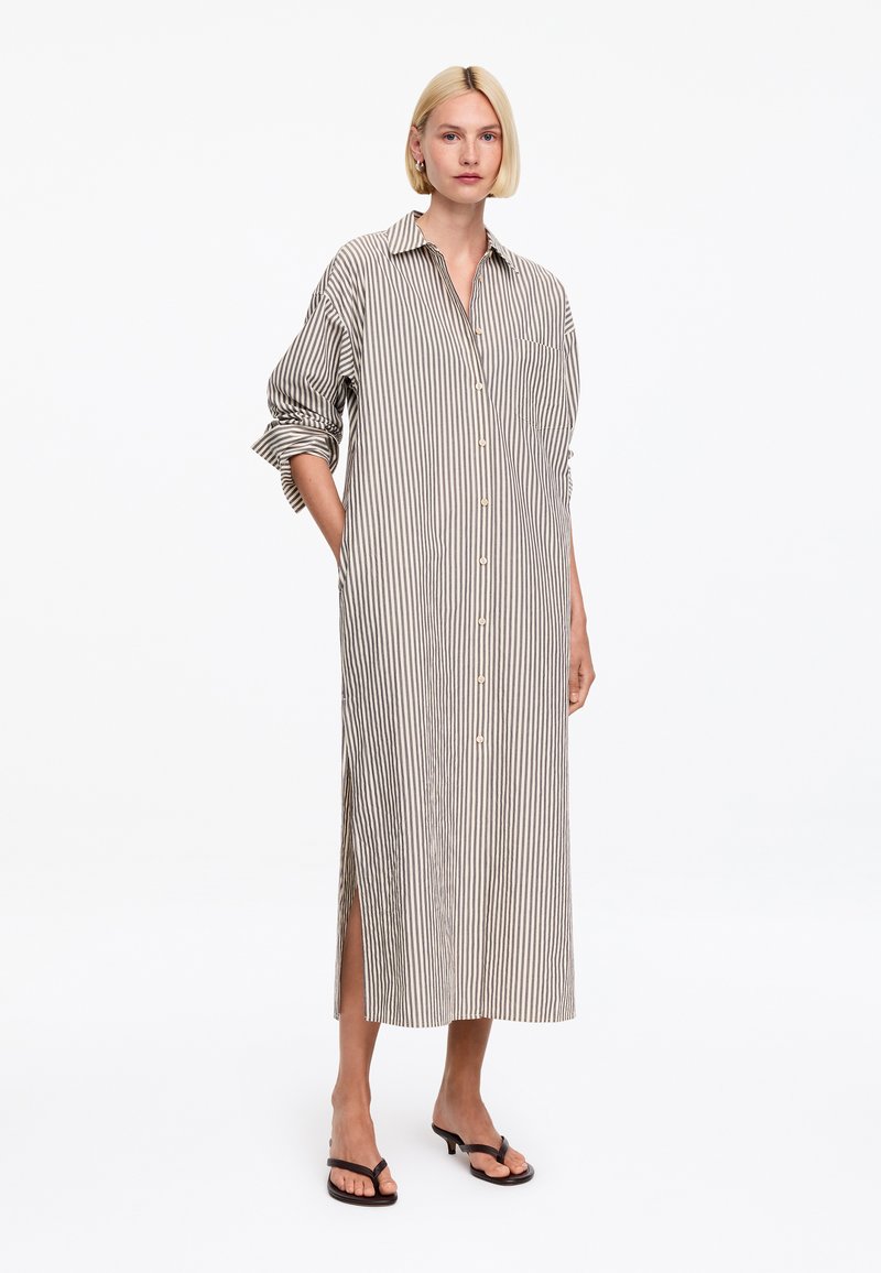 Femme portant une longue robe-chemise ample beige et blanche à rayures verticales, manches retroussées et sandales à talons noires.