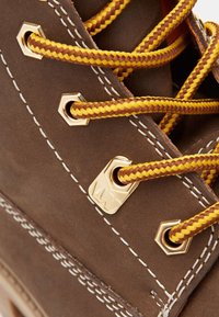 Bota de cuero marrón con ojales dorados, cordones trenzados en amarillo y marrón, y costuras blancas. La vista en primer plano resalta la textura y los detalles.