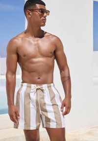 Beige und weiße gestreifte Badeshorts mit elastischem Bund und verstellbarem Kordelzug. Modell trägt Sonnenbrille und steht vor einem hellen Hintergrund.