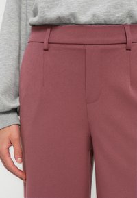 Mauve tailored trousers with a smooth, textured fabric. Features front pockets and a tailored waistband, paired with a gray sweater.