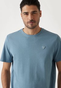 T-shirt in cotone azzurro chiaro con maniche corte, caratterizzato da un collo a giro e un piccolo logo a triangolo sul petto. Texture morbida e design semplice.