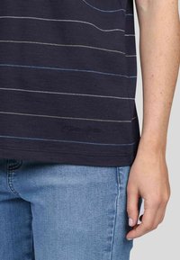 T-shirt rayé bleu marine avec des lignes de couleurs variées en bleu, vert et blanc ; texture douce ; logo brodé près de l'ourlet ; associé à un jean bleu clair.