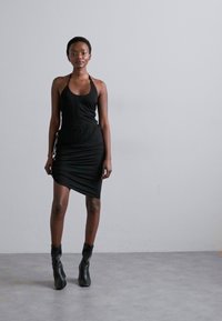 Rochie neagră fără mâneci, cu un design ajustat, având detalii pliate pe lateral. Purtați cu cizme negre până la gleznă, pe un fundal neutru.