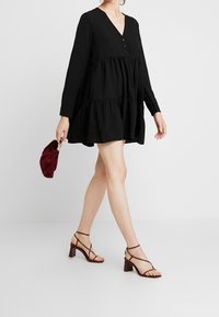Schwarzes schichtetes Kleid mit langen Ärmeln und V-Ausschnitt. Mittig mit Knopfdetails versehen. Dazu werden braune Riemchen-Highheels und eine kastanienbraune Clutch getragen.