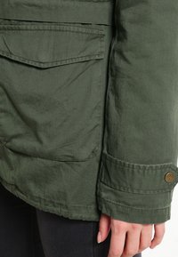 Chaqueta de algodón verde con un gran bolsillo parche, puño con botón y tejido texturizado. Detalles visibles de costuras y ligeras arrugas.