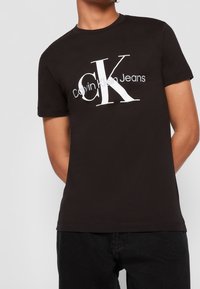Persona que lleva puesta una camiseta negra de Calvin Klein Jeans con un gran logo "cK" en blanco y gris en la parte delantera, combinada con pantalones negros.