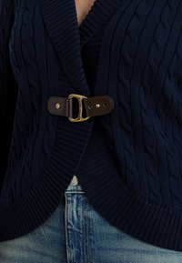 Tumši zils džempera stila kardigans ar adījumu un ribotām malām, pie jostas aizslēgts ar brūnu ādas sprādzi, valkāts pār zilām džinsa biksēm.