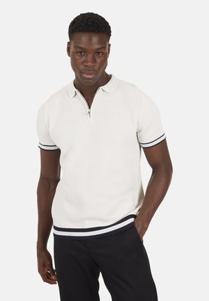 Giovane uomo con capelli corti e ricci che indossa una polo a maniche corte in maglia bianca con bordi a righe blu navy e azzurro, e pantaloni neri.