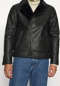 Czarna skórzana kurtka z sztucznym futrem przy kołnierzu, z zamkiem błyskawicznym z przodu i kieszeniami, o gładkiej fakturze w dopasowanym fasonie.