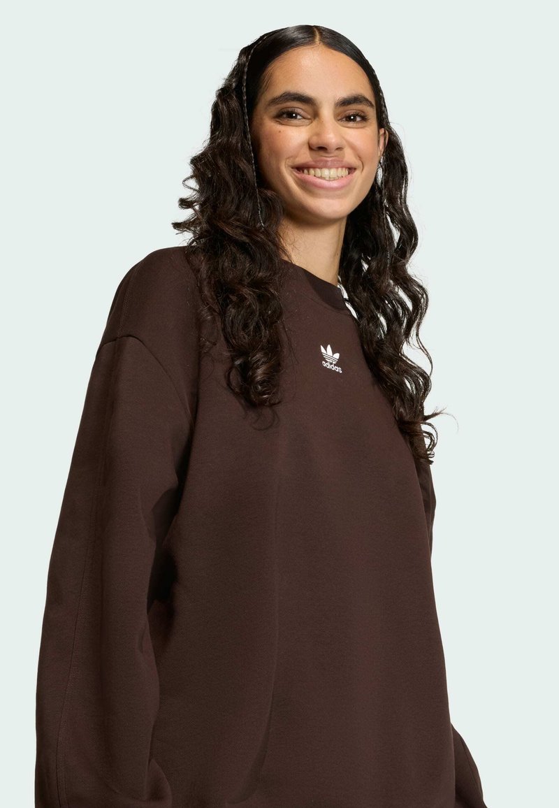 Woman with long curly hair wearing a brown Adidas sweatshirt, smiling gently, standing against a plain light background.