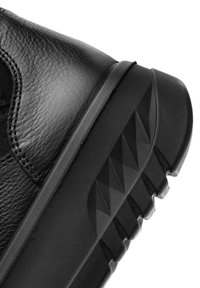 Zapatilla negra de cuero con una superficie texturizada, suela de goma robusta con un patrón de tracción triangular y detalles cosidos a lo largo de los bordes.