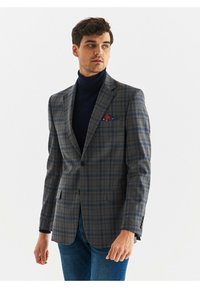 Szary w kratę żakiet wykonany z teksturowanej tkaniny, posiadający wyłogi, dwa guziki i niebieską kieszonkę w stylu poszetki z czerwonymi akcentami.