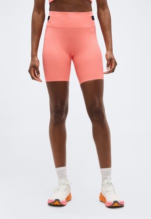 Mujer con shorts deportivos de talle alto en color coral, calcetines blancos y zapatillas blancas con suelas en naranja y rosa, de pie contra un fondo liso.