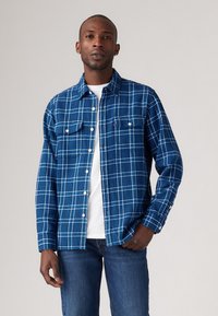 Chemise en flanelle à carreaux bleu avec un motif de carreaux bleu clair et blanc, deux poches poitrine, un bouton à l'avant et une coupe décontractée. Portée par-dessus un t-shirt blanc.