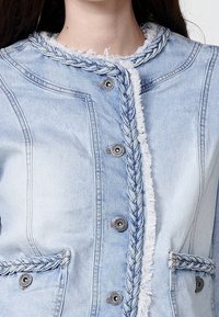 Light blue denim jacket with braided trim, frayed edges, and silver buttons. Features two front pockets and a structured design.