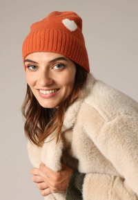 Oranje gebreide muts met ribbelstructuur en witte hartdetail, gedragen met een donzige beige jas, waardoor zachte materialen en contrasterende kleuren worden benadrukt.