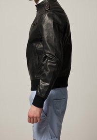 Svart läder bomberjacka med hög krage och tryckknapplås, ribbade ärmmuddar och sidofickor, tillsammans med ljusblå shorts.