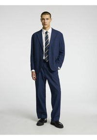 Costume bleu marine avec un blazer structuré et un pantalon à jambes larges. Comprend une chemise blanche et une cravate rayée. Les chaussures sont en cuir noir, brillant.
