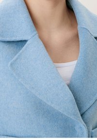 Ljusblå ullkappa med en mjuk textur, stor inskuren krage och knappstängning. Lager på en vit topp.