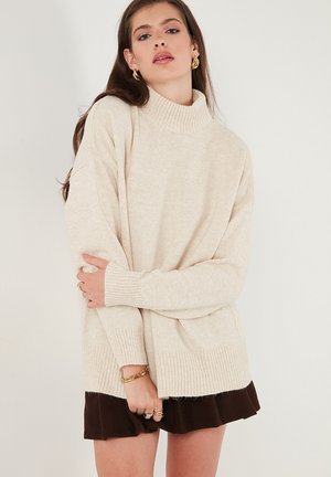 Maglione beige con collo alto, polsini e orlo a costine. Vestibilità oversize con una texture morbida. Abbinato a una gonna marrone.