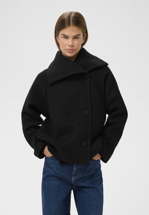 Sort uld-blandede cropped frakke med stor krave, tre knapper og sidelommer; parret med blå denim jeans. Minimalistisk design.