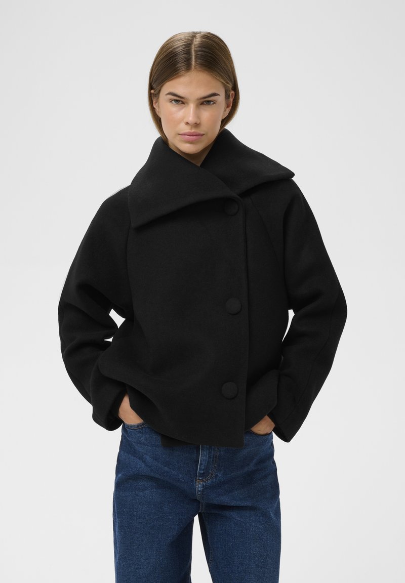 Sort uld-blandede cropped frakke med stor krave, tre knapper og sidelommer; parret med blå denim jeans. Minimalistisk design.